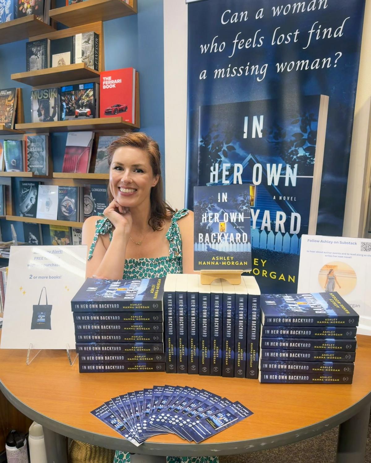 Ashley Hanna-Morgan smiling at a round signing table at Warwick’s in La Jolla, stacked with copies of In Her Own Backyard and fanned bookmarks, in front of a tall banner of the book’s cover asking “Can a woman who feels lost find a missing woman?”