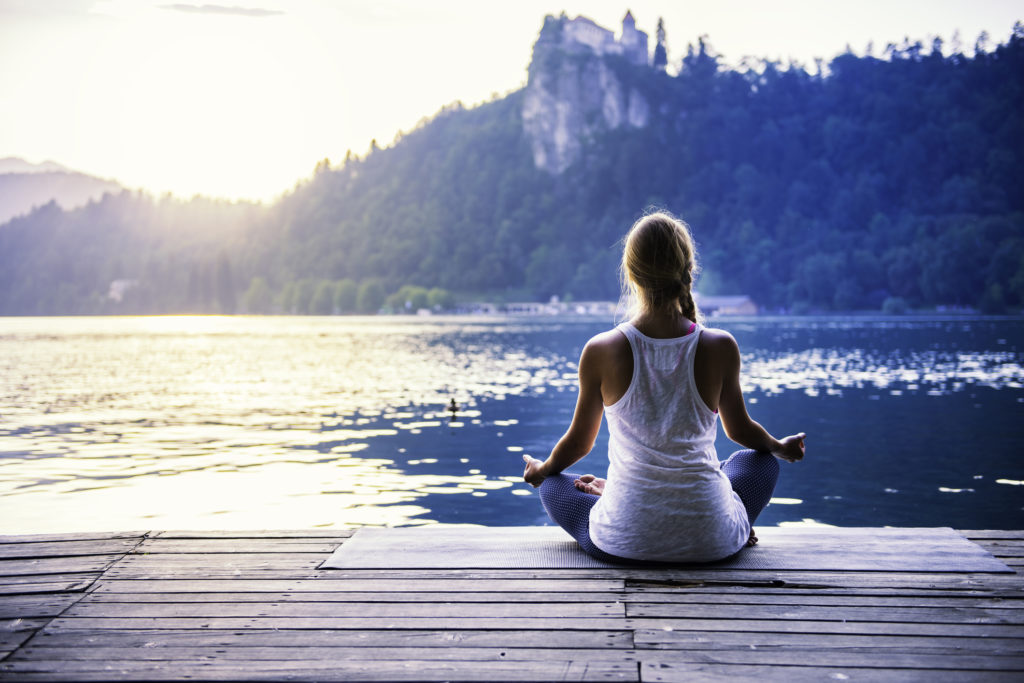 A woman sitting cross-legged on a wooden dock, facing a calm lake and mountains.